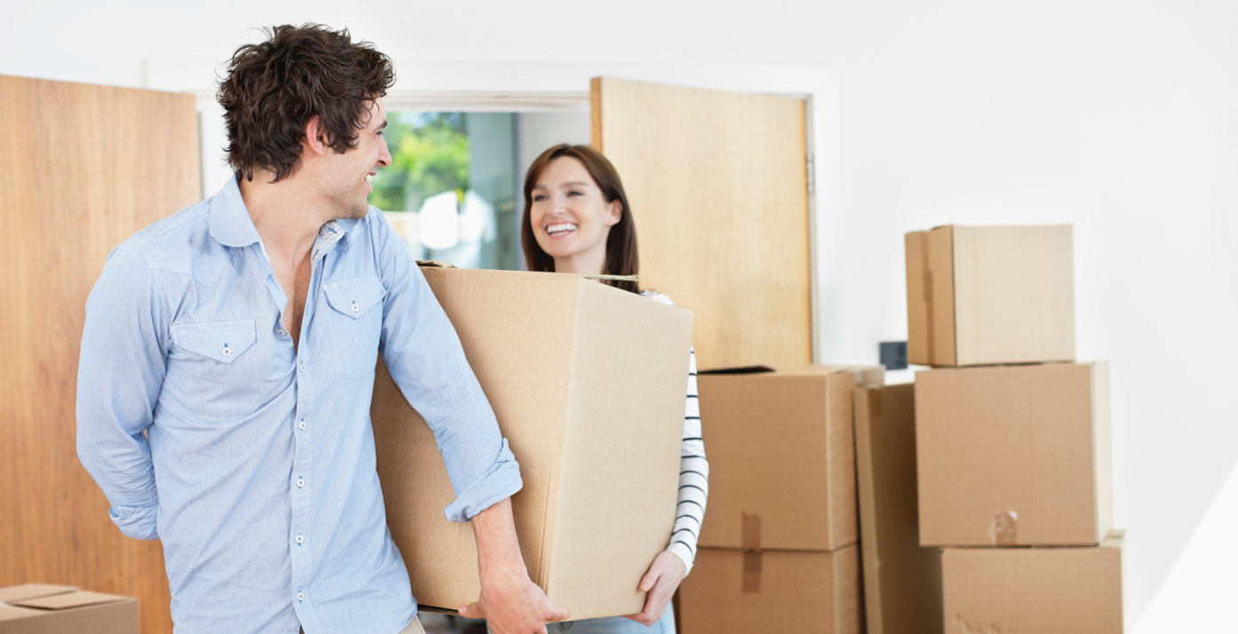 Couple smiling at each other carrying a moving box with boxes in the background